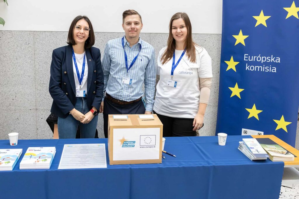 Tri osoby stoja za stolom s brožúrami, papiermi, kartónom a materiálmi na Deň peňazí. Vedľa nich je vyvesený transparent s hviezdami EÚ a Európskou komisiou. Všetci sa usmievajú a majú na sebe menovky.