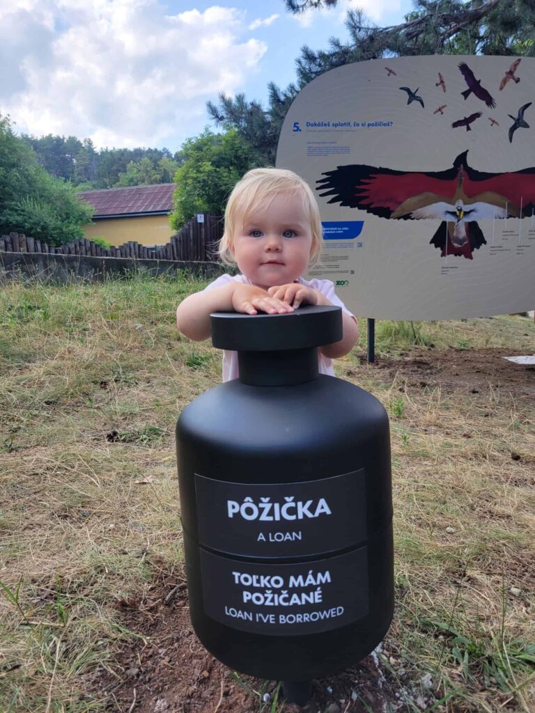 Malé dieťa stojí vonku a drží veľký čierny valec s nápisom PÔŽIČKA / A LOAN / TOĽKO MÁM POŽIČANÉ / LOAN IVE BORROWED. V pozadí sa nachádza vývesná tabuľa s ilustráciou vtáka.