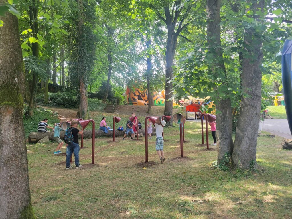 Deti sa hrajú vonku v zalesnenom parku a používajú červené a strieborné hovoriace trubice. Stromy poskytujú tieň a v pozadí je farebná nafukovacia konštrukcia s ľuďmi v blízkosti. Slnečné svetlo preniká cez listy.