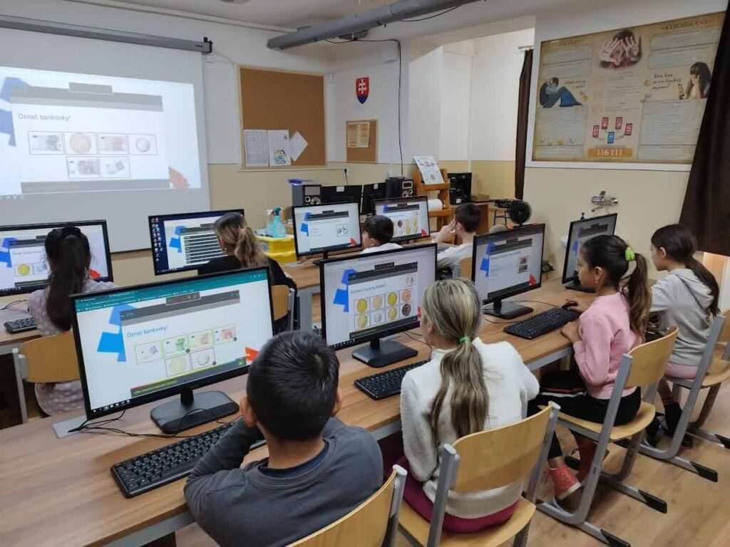 Deti sedia za stolmi v počítačovej učebni a každý používa stolný počítač. Vpredu stojí učiteľ a na obrazovke sa premieta učivo. Na stenách visia vzdelávacie plagáty a tabuľa.