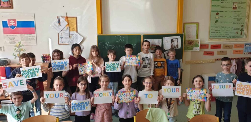 Skupina školákov stojí v triede a drží v rukách nápisy: SAVE YOUR MONEY TO YOUR FUTURE (Šetrite si peniaze na svoju budúcnosť). Na niektorých z nich je napísané aj Global Money Week. Deti sa usmievajú a za nimi je tabuľa.