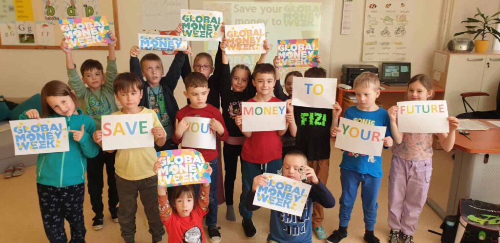 Skupina detí v triede drží farebné nápisy GLOBAL MONEY WEEK a SAVE YOUR MONEY TO YOUR FUTURE. Usmievajú sa a stoja spolu pred tabuľou.