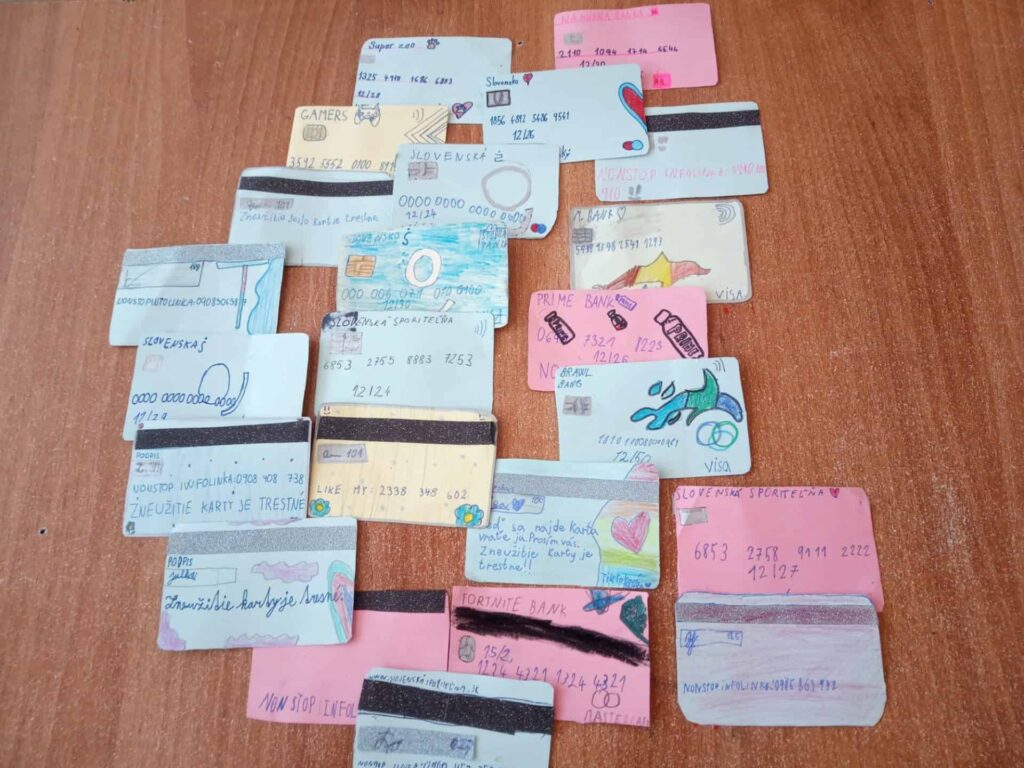 Na drevenom povrchu je rozmiestnených niekoľko ručne kreslených farebných imitácií bankových kariet s ručne písanými detailmi a čmáranicami. Každá karta je jedinečná, obsahuje čiarové kódy, čísla a kreatívne ilustrácie.