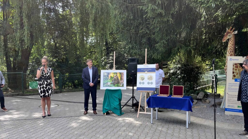 Žena hovorí do mikrofónu vedľa muža v obleku na podujatí pod holým nebom. Neďaleko sú výstavné tabule, plakety na stole a socha žirafy, v pozadí sú stromy a slnečné svetlo.
