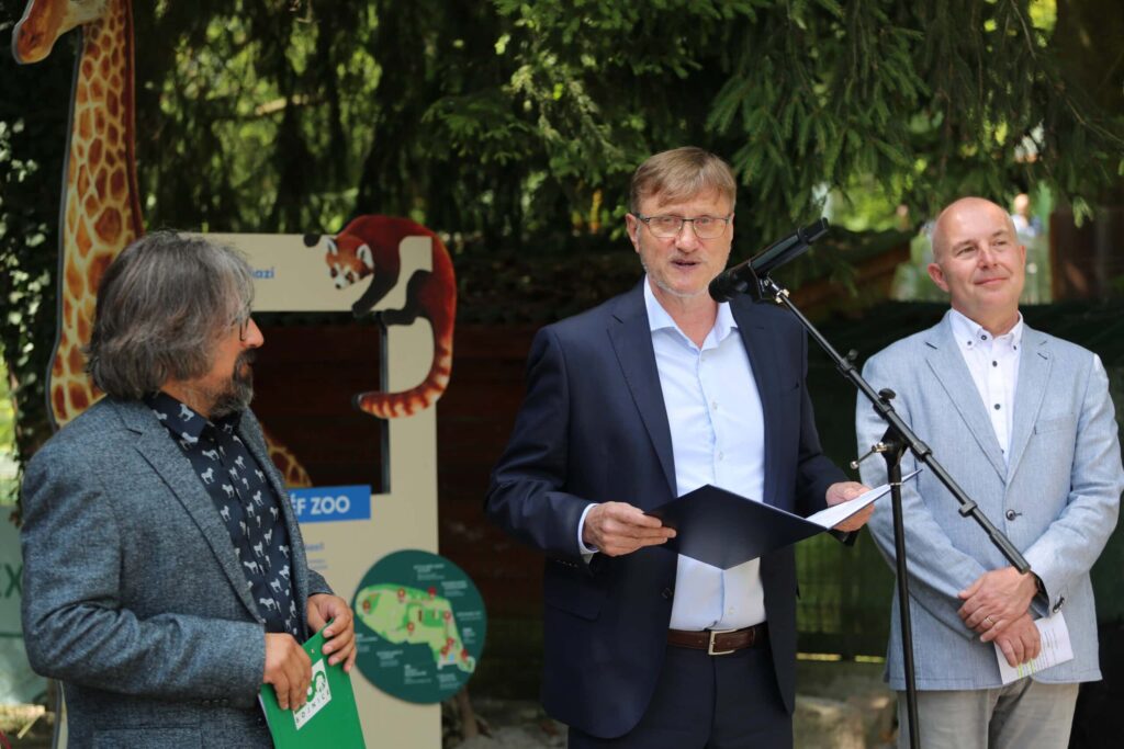 Traja muži stoja vonku pred mikrofónom. Jeden muž v tmavom obleku hovorí a drží v ruke papiere; ostatní počúvajú. V pozadí je vidieť výrez žirafy a nápis zoologickej záhrady. Skupinu obklopujú stromy.