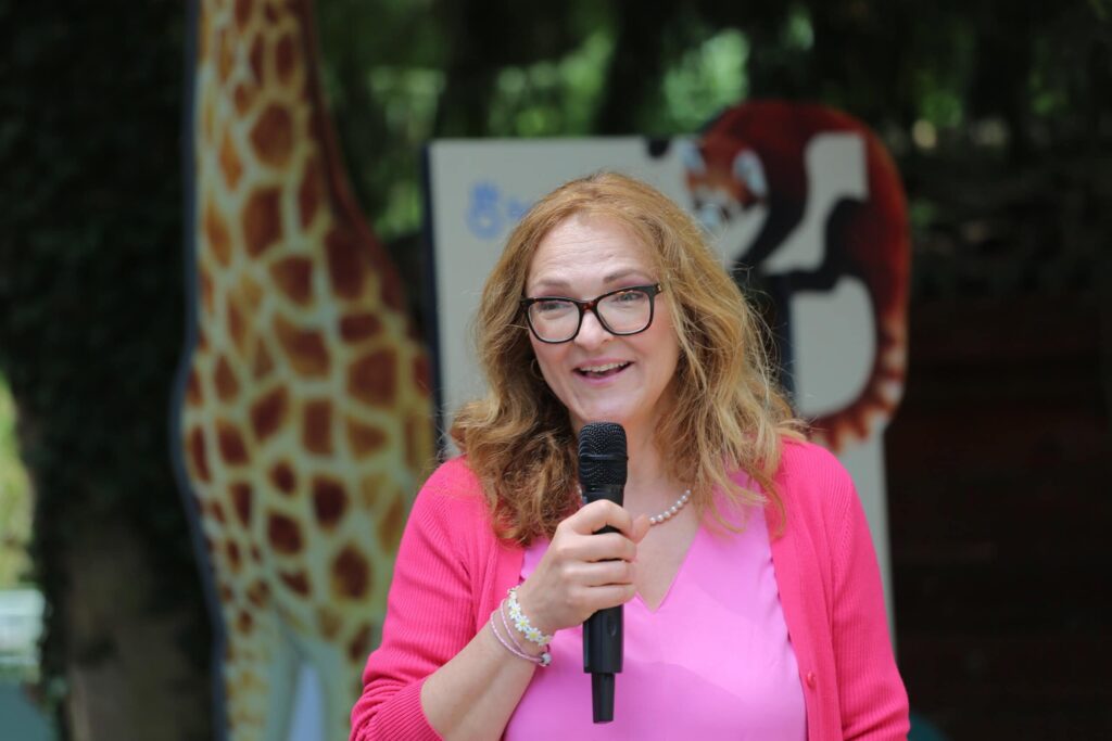Žena s vlnitými blond vlasmi, okuliarmi a ružovým oblečením sa usmieva a drží mikrofón. V pozadí sú obrázky žirafy a červeného zvieraťa, ktoré sú pravdepodobne súčasťou zoologickej záhrady alebo vzdelávacieho zariadenia.