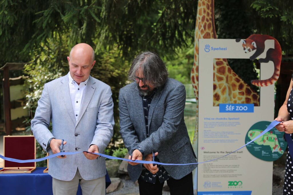 Dvaja muži v oblekoch prestrihávajú modrú pásku na podujatí pod holým nebom, v pozadí sú stromy a informačná tabuľa zoologickej záhrady so žirafou a pandou červenou. Vpravo je vidieť ženskú ruku, ktorá drží stuhu.