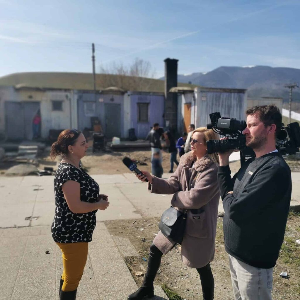 Reportér s mikrofónom v ruke robí rozhovor so ženou, zatiaľ čo ich kameraman natáča. V pozadí je niekoľko ľudí a schátraná budova, v diaľke vidno hory.