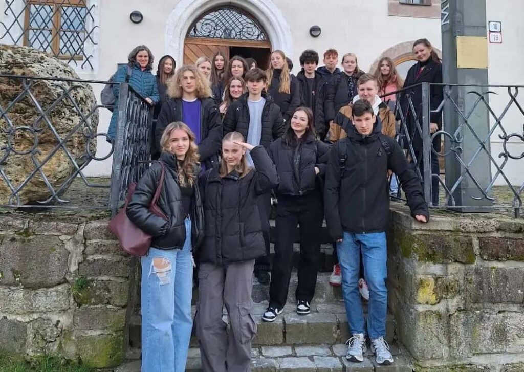 Skupina tínedžerov a jeden dospelý stoja na kamenných schodoch pred budovou s klenutými drevenými dverami a železným zábradlím a pózujú na skupinovú fotografiu. Väčšina z nich má na sebe bundy a bežné oblečenie.