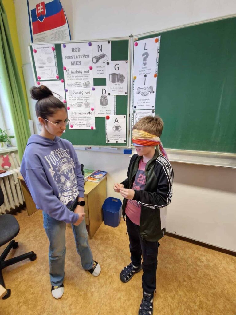 Dve deti stoja v triede; jeden chlapec má zaviazané oči a drží nejaký predmet, zatiaľ čo druhé dievča ho pozoruje. V pozadí sú viditeľné vzdelávacie plagáty a tabuľa.