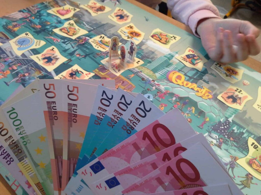 Detailný záber na stolnú hru s farebnou hracou doskou, hernými figúrkami a vejárom eurobankoviek. Vidno detskú ruku, čo naznačuje aktívnu hru.