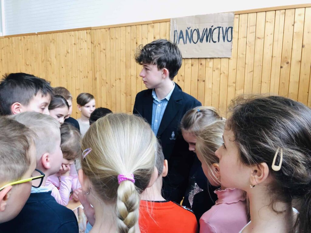 Skupina detí sa zhromažďuje okolo chlapca v saku, ktorý stojí uprostred. Zdá sa, že prostredie je v interiéri s dreveným obložením a ručne písaným nápisom v pozadí.
