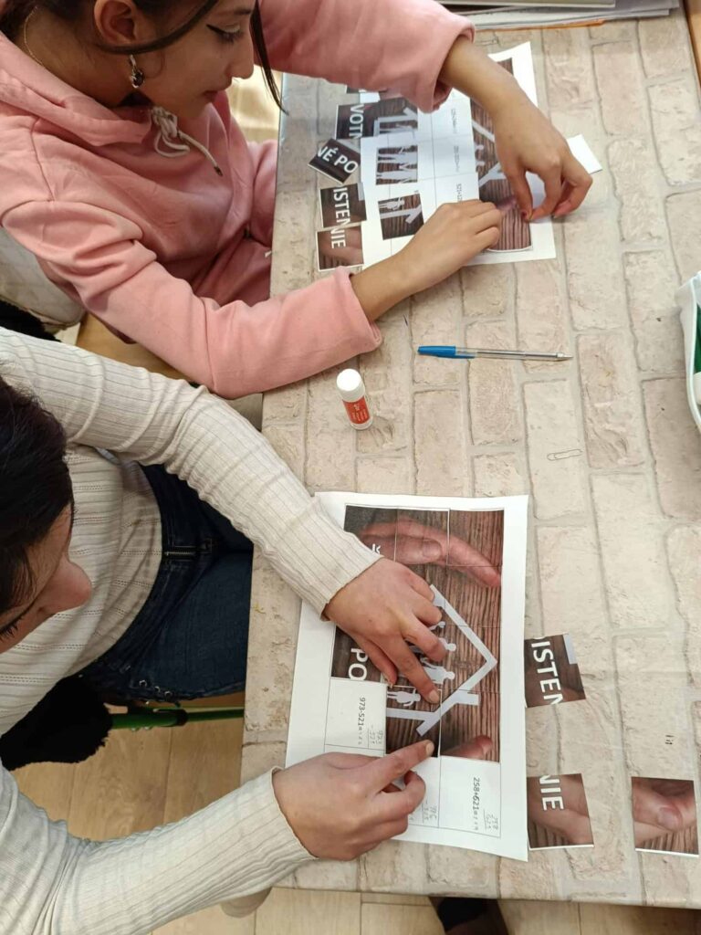 Dvaja ľudia sedia za stolom a pracujú na papierovej remeselnej činnosti, pričom pomocou lepiacich tyčiniek skladajú vytlačené kúsky skladačky na hárky papiera. Na stole je svetlý tehlový vzor.