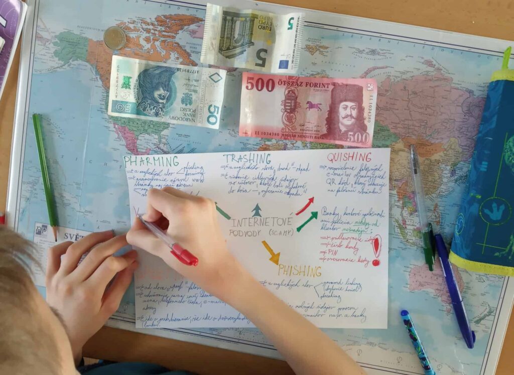 Dieťa píše na farebný plagát o bezpečnosti na internete o phishingu a podvodoch, obklopené perami, ceruzkou, rôznymi bankovkami a mapou sveta na stole.