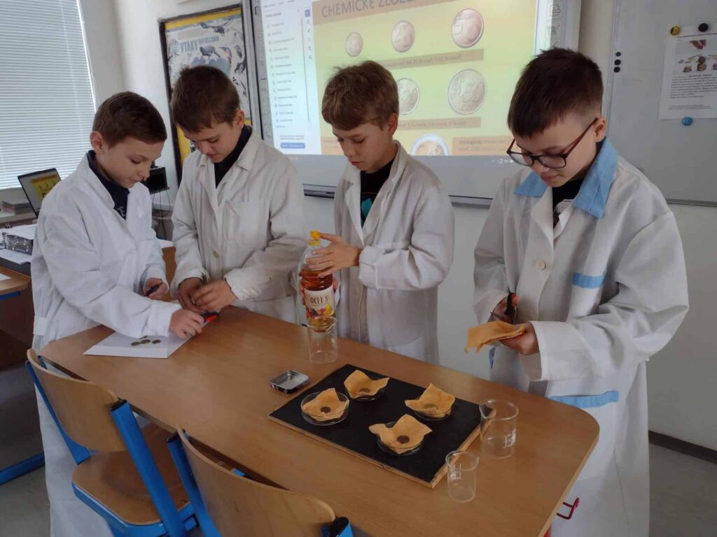 Štyria chlapci v laboratórnych plášťoch stoja okolo stola v triede a vykonávajú pokus s mincami, kadičkami, handričkami a fľašou s octom, zatiaľ čo na obrazovke v pozadí sa zobrazuje prezentácia.