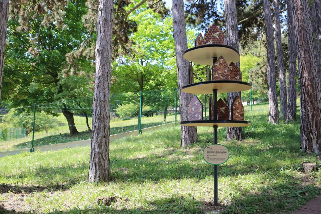 Moderný hotel pre hmyz s kruhovými vrstvenými plošinami stojí medzi vysokými stromami v trávnatom parku. Slnečné svetlo preniká cez listy a v pozadí sa tiahne zelený plot.