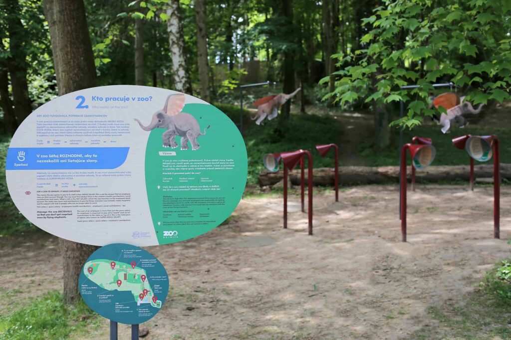Náučná tabuľa zoologickej záhrady s ilustráciami slona a žirafy stojí v zalesnenej hracej ploche so stavbami tunelov a veľkými výrezmi dinosaurov v pozadí. Zem je piesočnatá a priestor obklopujú stromy.