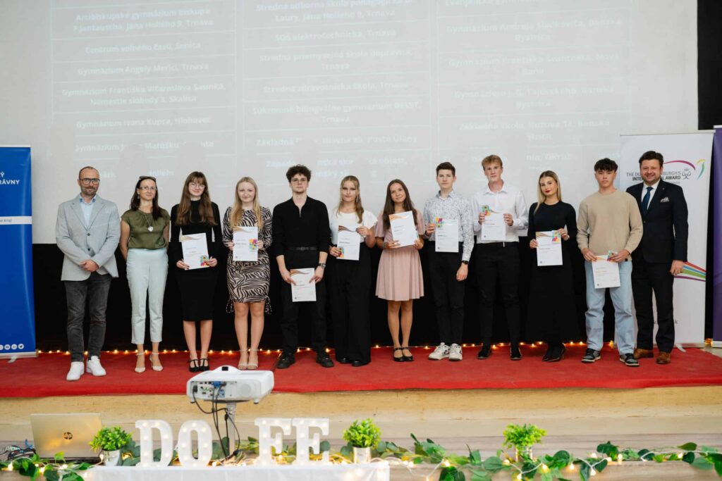 Skupina jedenástich mladých ľudí a dvoch starších ľudí stojí na pódiu a drží certifikáty. Sú formálne oblečení a pózujú na fotografiu pred premietacím plátnom. Pódium je vyzdobené zeleňou a veľkými písmenami s nápisom DOFE.