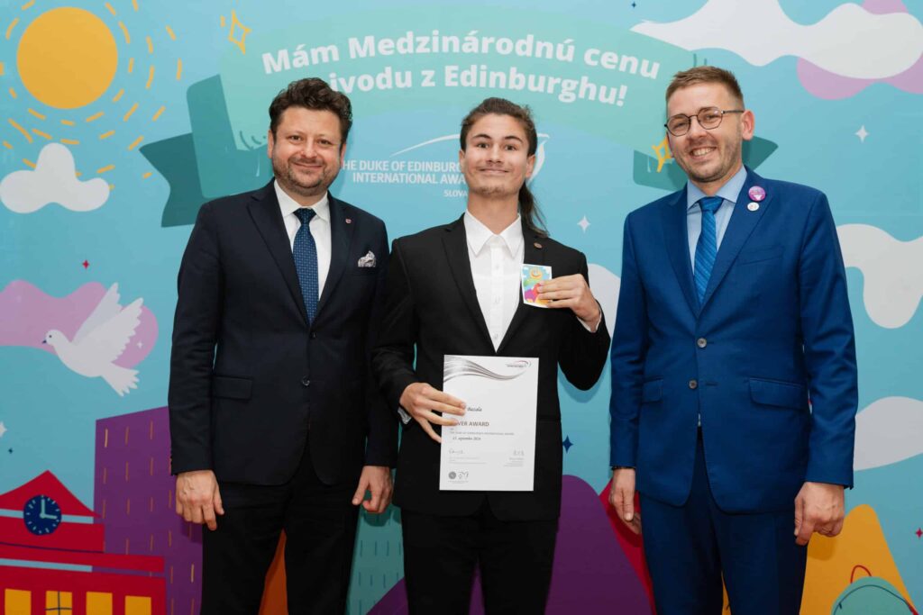 Traja muži v oblekoch stoja s úsmevom pred farebným pozadím. Muž uprostred drží certifikát a odznak a oslavuje úspech. Na pozadí je slovenský text a veselé ilustrácie.