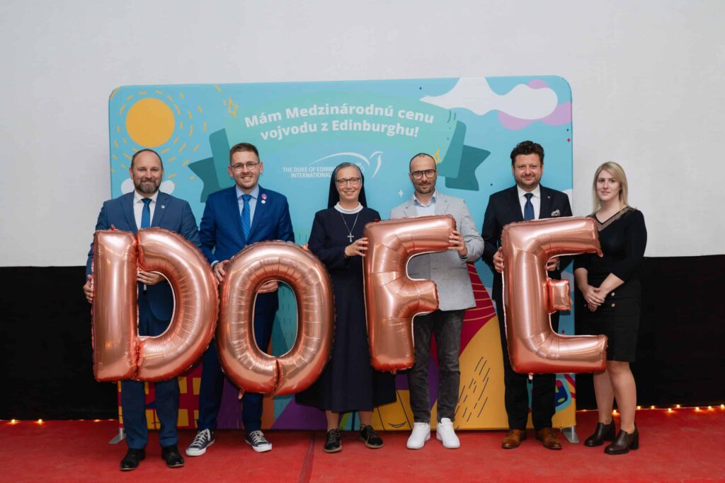 Šesť dospelých stojí na červenom koberci a drží veľké balónové písmená s nápisom DOFE pred farebným pozadím so slovenským textom a grafikou. Všetci sa usmievajú a sú formálne oblečení.