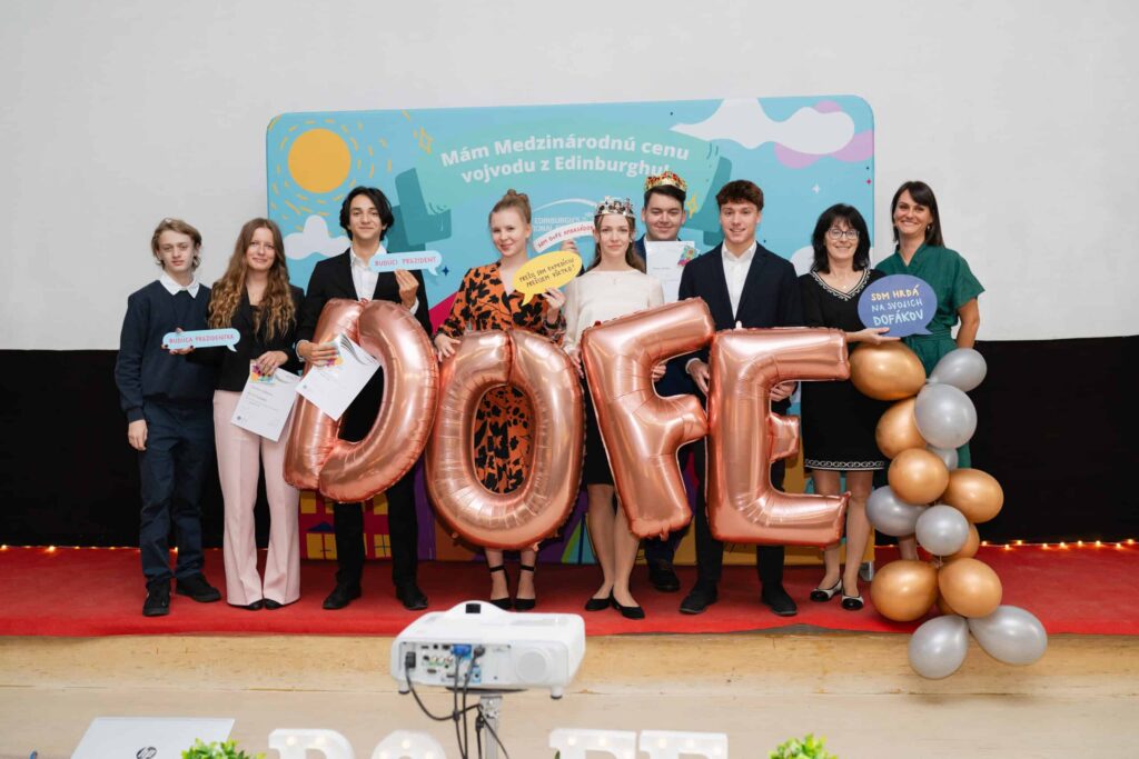 Skupina mladých ľudí stojí na pódiu s veľkými kovovými balónmi a certifikátmi "DOFE" a usmieva sa do kamery. Po oboch stranách stoja dvaja dospelí, za ktorými je vyzdobené pozadie.