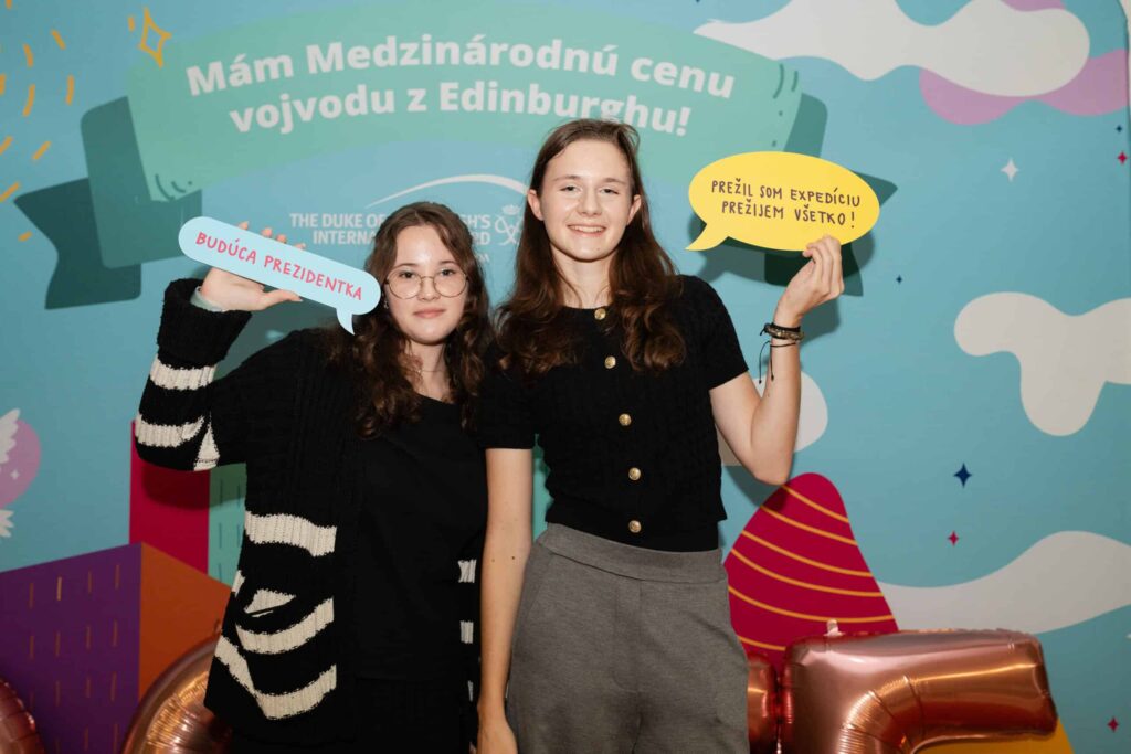 Dve mladé ženy stoja s úsmevom pred farebným pozadím so slovenským textom. V rukách držia ozdobné rečové bubliny; na jednej je napísané BUDÚCA PREZIDENTKA a na druhej PREŽIJEM EXPEDÍCIU, PREŽIJEM VŠETKO!.