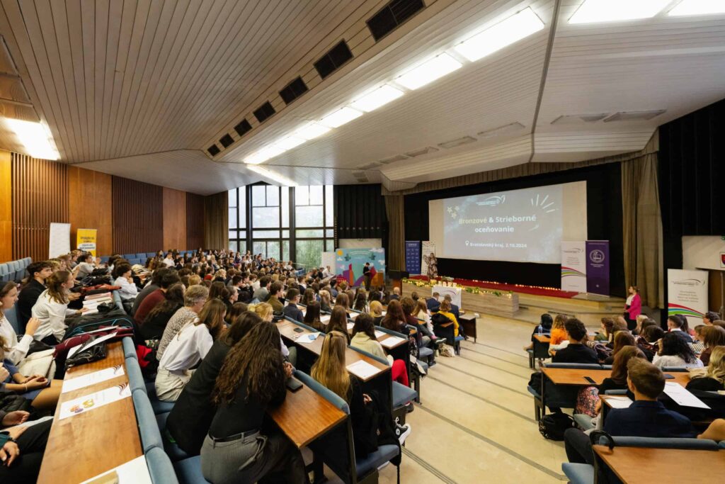 Veľká prednášková sála plná študentov sediacich v laviciach pred pódiom s reproduktormi a veľkým premietacím plátnom, na ktorom sa zobrazuje prezentácia. Prirodzené svetlo vchádza cez vysoké okná na ľavej strane.