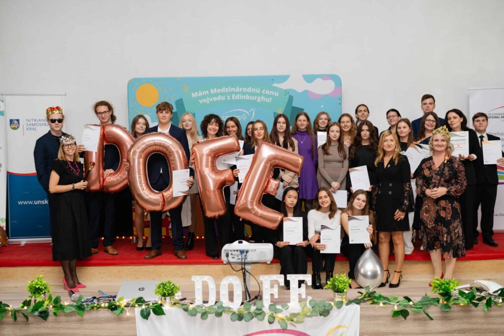 Veľká skupina ľudí, väčšinou mladých dospelých, pózuje na pódiu s certifikátmi. Štyri osoby vpredu držia veľké balónové písmená s nápisom "DOFE". Na pozadí sú farebné dekorácie a transparenty.