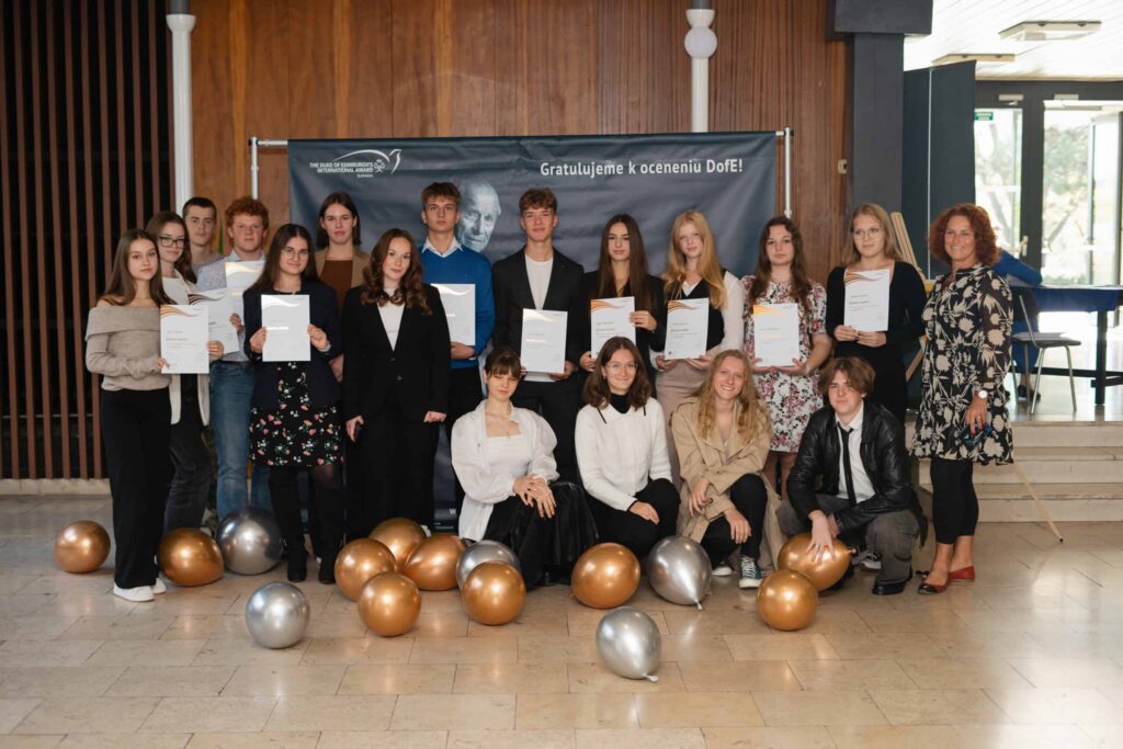 Skupina mladých ľudí a dvaja dospelí stoja v interiéri a pózujú na fotografiu s certifikátmi. Na zemi pred nimi ležia zlaté a strieborné balóny a v pozadí visí blahoprajný transparent.