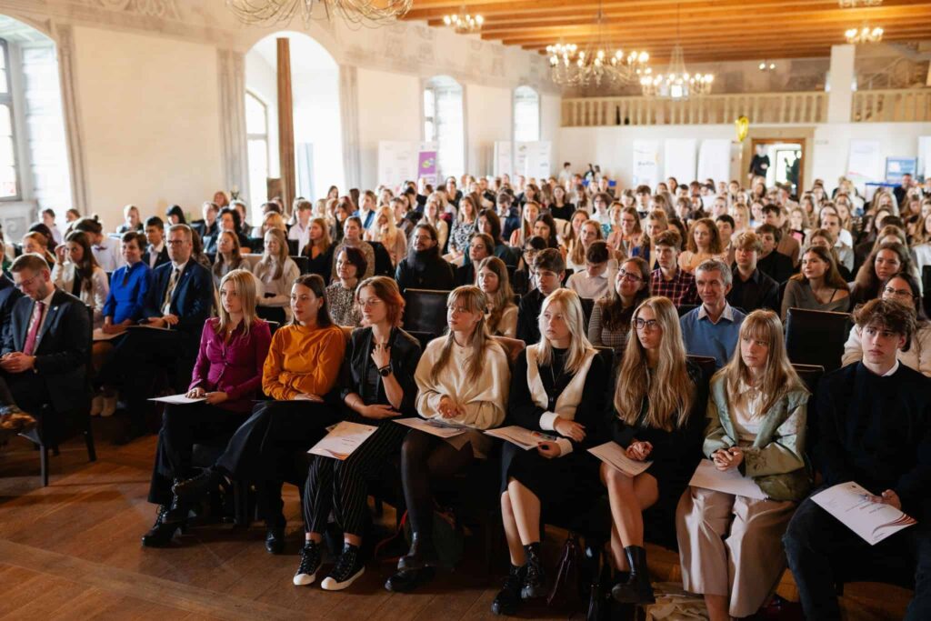 Veľká skupina ľudí, väčšinou mladých dospelých, sedí pozorne v radoch stoličiek na podujatí alebo konferencii v interiéri v priestrannej sále s drevenými podlahami, vysokými stropmi a lustrami.