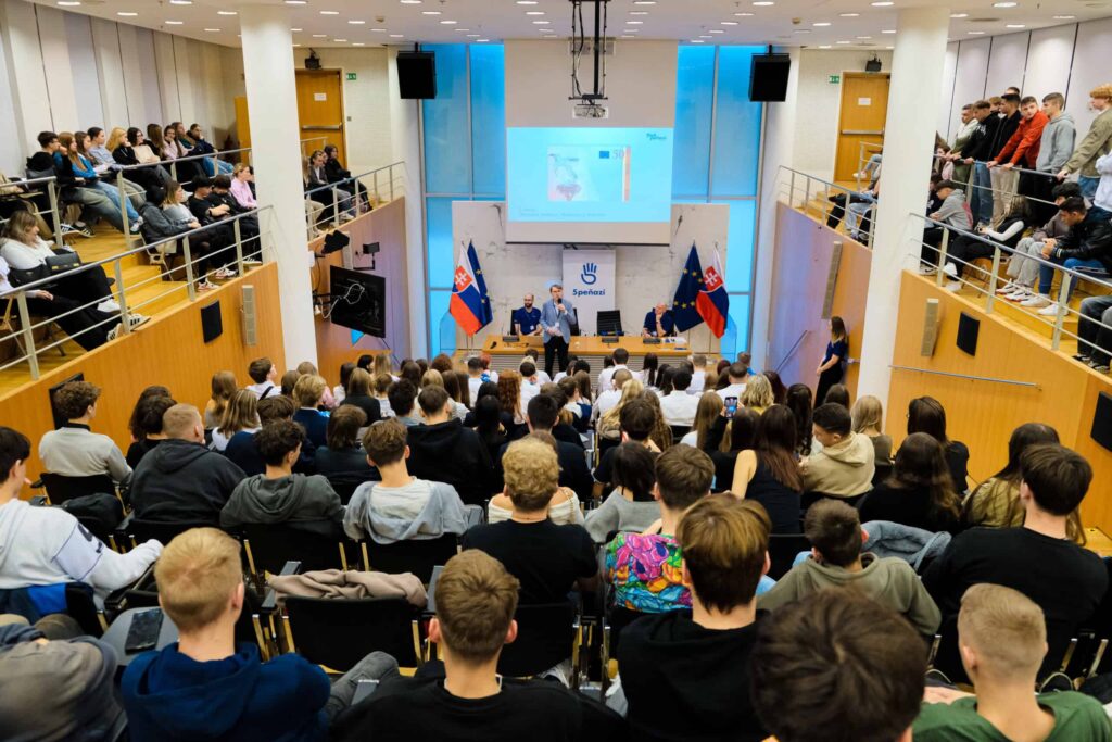 Veľká skupina ľudí sa zúčastňuje na konferencii alebo prednáške v modernej posluchárni, vpredu sedia rečníci, na plátne je prezentácia a po oboch stranách sú vyvesené slovenské vlajky.
