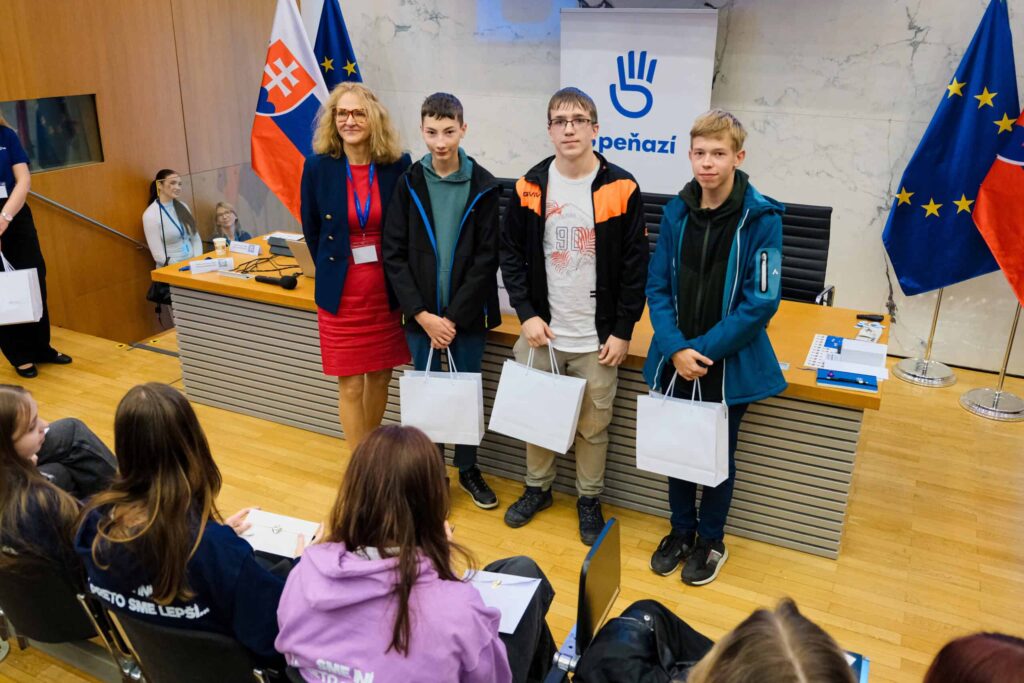 Pred stolom stoja traja chlapci s taškami na darčeky a usmievajú sa vedľa ženy v červených šatách a saku. Scéna sa odohráva v konferenčnej miestnosti, na ktorej sú viditeľné slovenské vlajky a vlajky Európskej únie. V popredí sedia diváci.