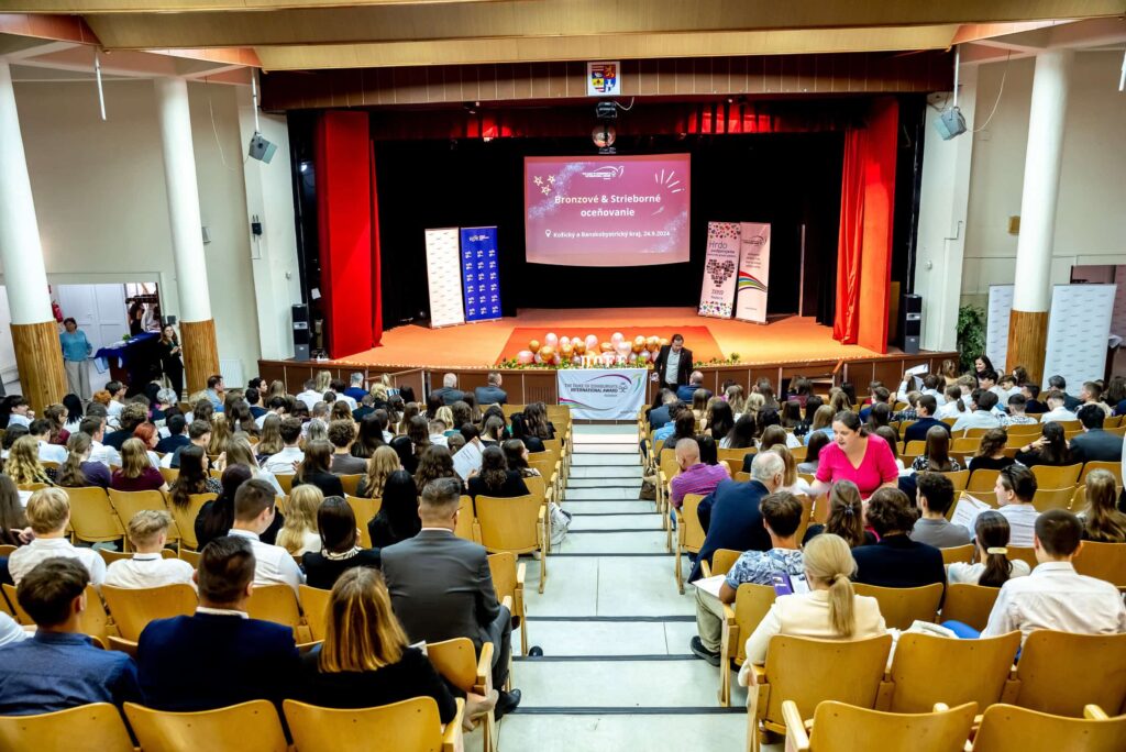Početné publikum sedí v radoch sály, pred pódiom s červenou oponou, premietacím plátnom s farebnou grafikou a niekoľkými stolmi ozdobenými balónmi. Vpredu stojí rečník, ktorý sa prihovára davu.