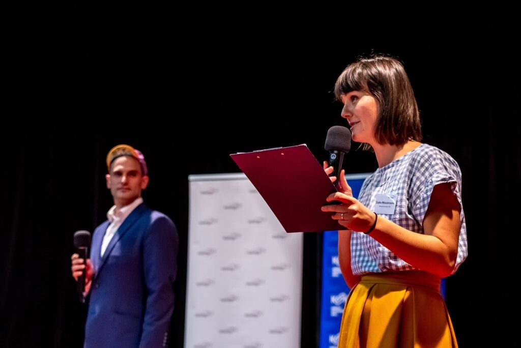 Na pódiu hovorí do mikrofónu žena s klipom v ruke a v pozadí stojí muž v obleku a čiapke, ktorý tiež drží mikrofón, a za nimi je tmavé pozadie.