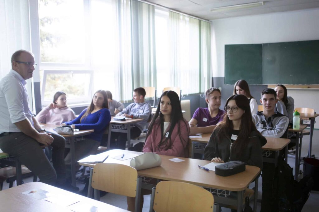 Učiteľ sedí na lavici vpredu v triede a hovorí k skupine pozorných stredoškolákov, ktorí sedia v laviciach so zošitmi a taškami pred sebou. V miestnosti sú svetlé okná a tabuľa.
