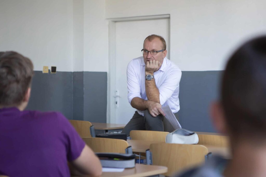 Muž v bielej košeli a okuliaroch sedí na lavici v prednej časti triedy, nakláňa sa dopredu, bradu si opiera o ruku a zamyslene sa pozerá na dvoch študentov v popredí.