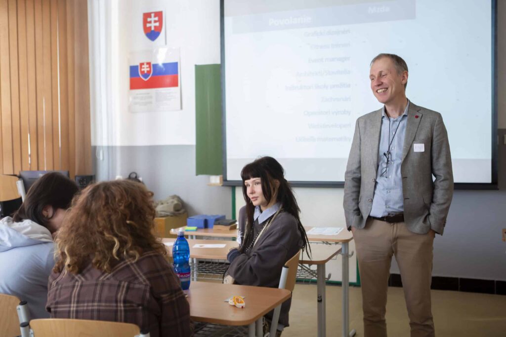 Muž v obleku stojí a usmieva sa počas rozhovoru so skupinou študentov sediacich v laviciach v triede. V pozadí je vidieť premietacie plátno, slovenské vlajky a učebné pomôcky.