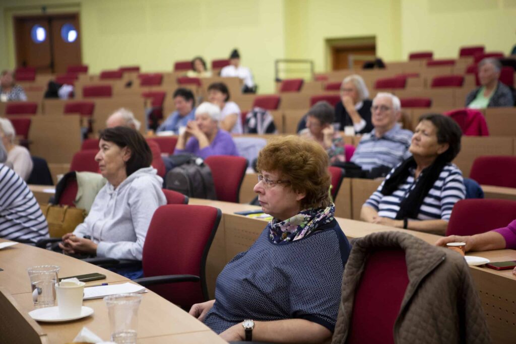 Skupina dospelých, väčšinou seniorov, sedí pozorne v prednáškovej sále s červenými stoličkami a drevenými stolmi a počúva rečníka mimo záberu. Niektorí majú na stoloch notebooky a nápoje.