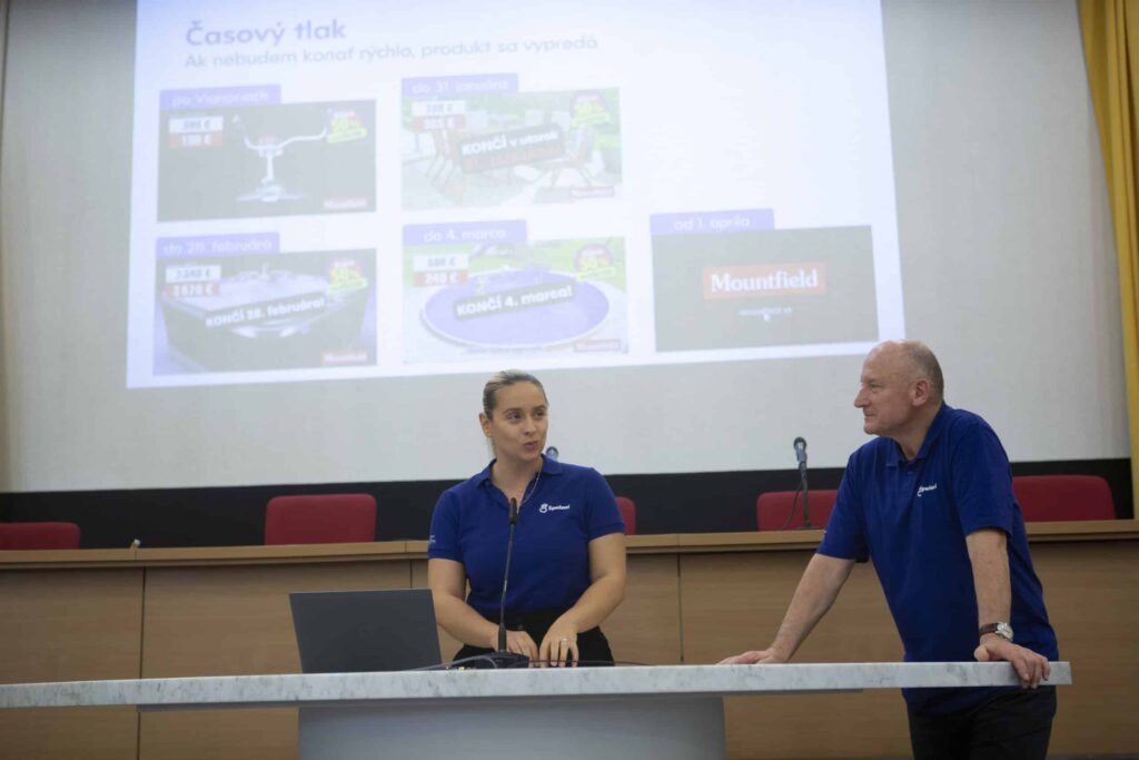 Dvaja ľudia v modrých košeliach stoja na pódiu v konferenčnej miestnosti a za nimi sa na veľkej obrazovke zobrazuje prezentácia v slovenčine. Žena hovorí, zatiaľ čo muž počúva.
