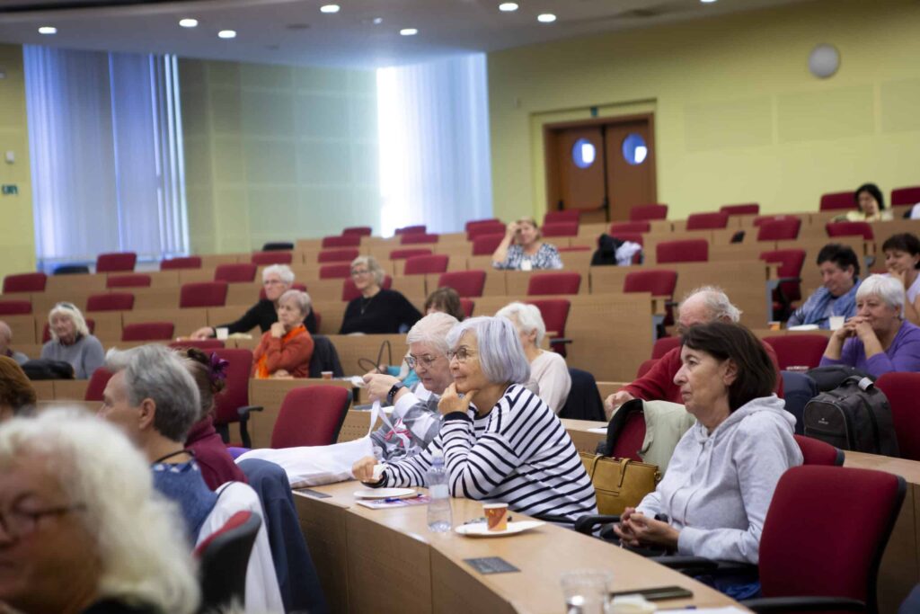 Skupina starších ľudí sedí v stupňovitej prednáškovej sále a pozorne počúva prezentáciu. Niektorí si robia poznámky, zatiaľ čo iní sledujú prednášajúceho. V miestnosti sú červené stoličky a veľké okná prepúšťajúce prirodzené svetlo.
