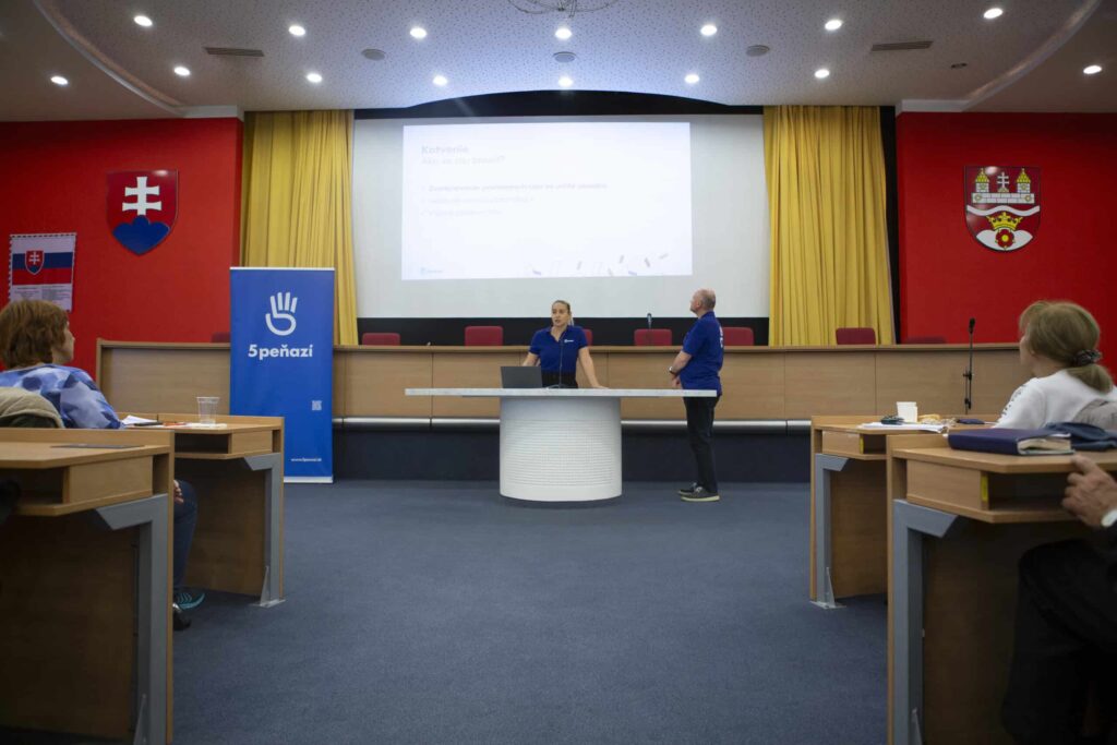 Dvaja ľudia stoja na pódiu a prezentujú publiku v konferenčnej miestnosti. Na veľkej obrazovke sa zobrazuje prezentácia. Na červených stenách za nimi sú viditeľné slovenské vlajky a transparenty.