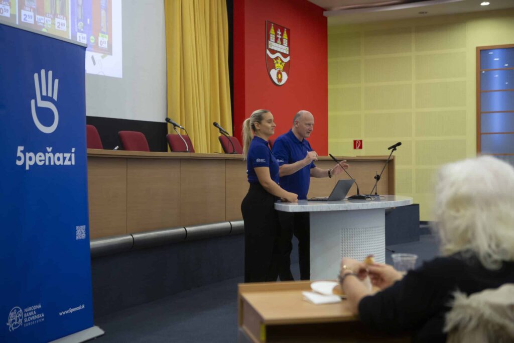 Dvaja prednášajúci v modrých košeliach stoja na pódiu v konferenčnej miestnosti a prihovárajú sa publiku. Naľavo je vidieť veľký modrý transparent s nápisom "5PEŇAZÍ" a logom ruky. V popredí sedí žena a pozoruje rečníkov.