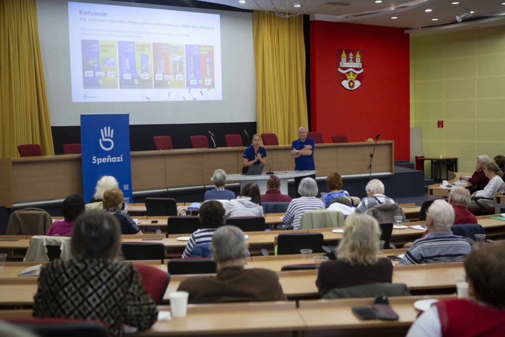 Žena a muž prednášajú poslucháčom, ktorí sedia v prednáškovej sále, na plátne sa premietajú diapozitívy a vpredu v miestnosti je modrý transparent s ikonou ruky.