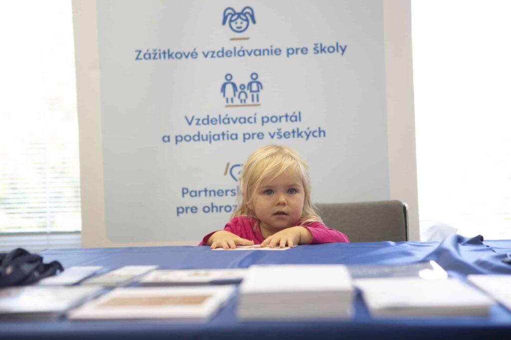 Mladé dievča so svetlými vlasmi sedí za stolom pokrytým brožúrami a papiermi. Za ňou je veľký informačný banner so slovenským textom a vzdelávacími ikonami.