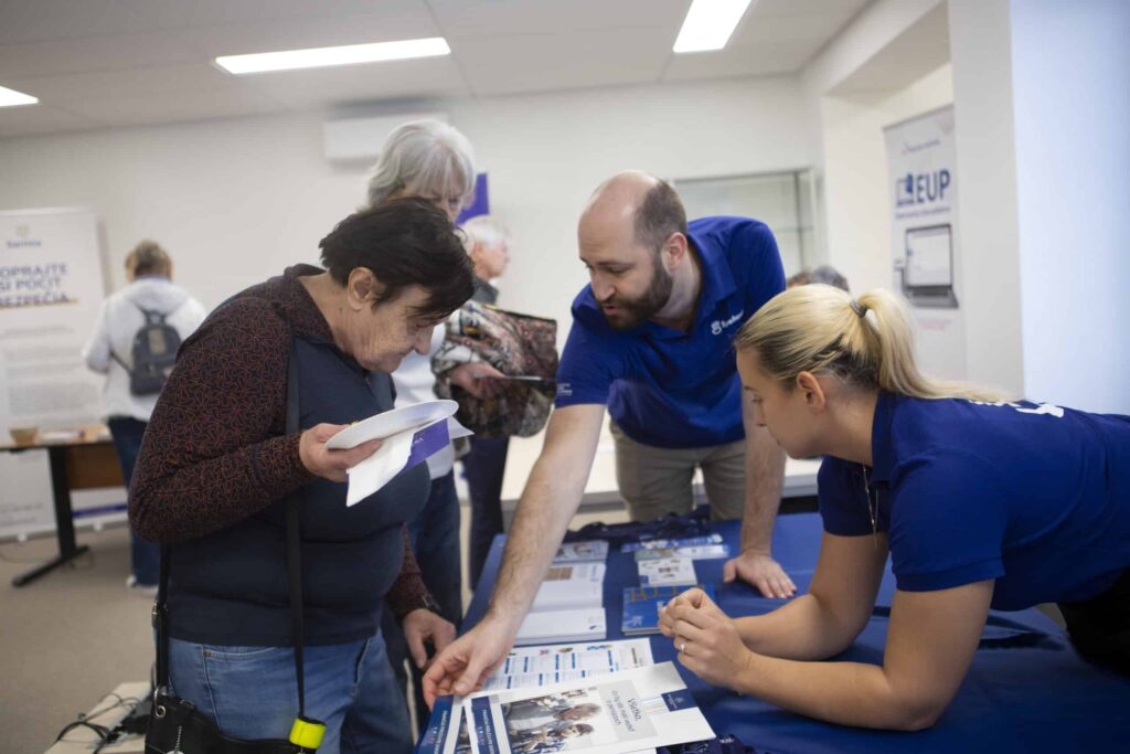 Muž a žena v modrých košeliach pomáhajú staršej žene pri informačnom stánku a ukazujú na letáky na stole. Zdá sa, že ide o komunitné alebo informačné podujatie v interiéri.