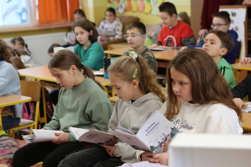 Trieda plná študentov sedí v laviciach, niektorí čítajú z brožúr a iní sa pozorne pozerajú dopredu. Trieda je svetlá, s farebnými dekoráciami a oranžovými závesmi na oknách.