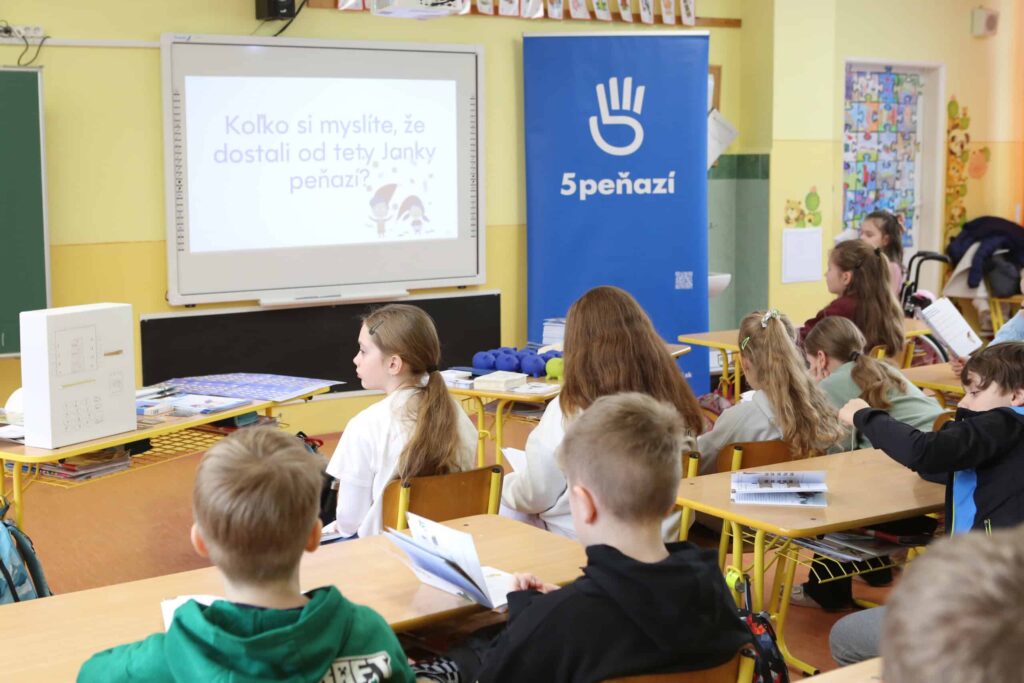 Trieda so študentmi sediacimi v laviciach, ktorí stoja pred premietacím plátnom s otázkami v slovenčine. Pri plátne stojí modrý transparent s nápisom 5PEŇAZÍ a symbolom bielej ruky. Žiaci držia v rukách zošity a vyzerajú pozorne.
