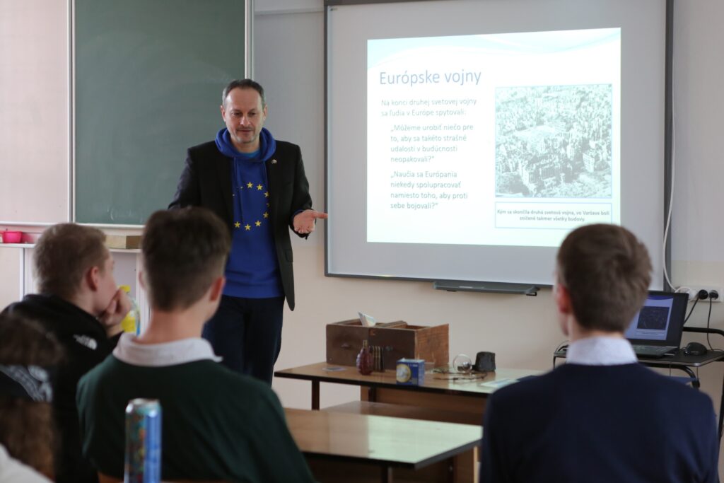 Muž v modrej mikine s kapucňou EÚ sa prihovára študentom v triede. Projektor zobrazuje diapozitív s názvom Európske vojny s textom a čiernobielou fotografiou. Štyria študenti stoja tvárou k prednášajúcemu.