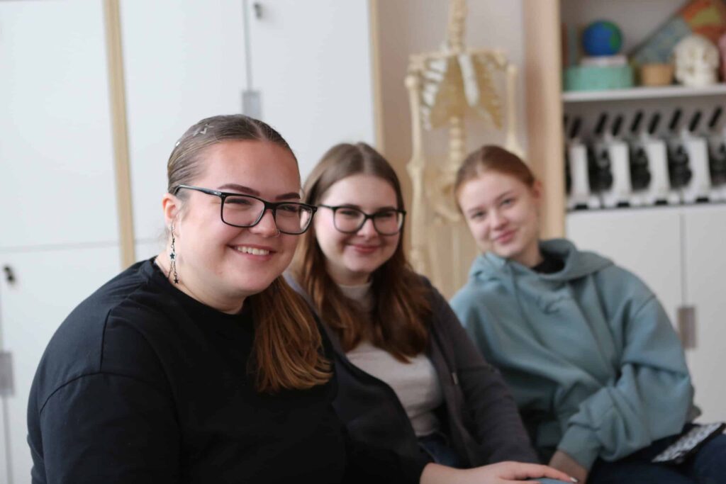 Tri mladé ženy s dlhými vlasmi a okuliarmi sa usmievajú a sedia spolu vo vnútri. V pozadí je viditeľný model ľudskej kostry a police s učebnými pomôckami, čo naznačuje prostredie triedy alebo učebne.