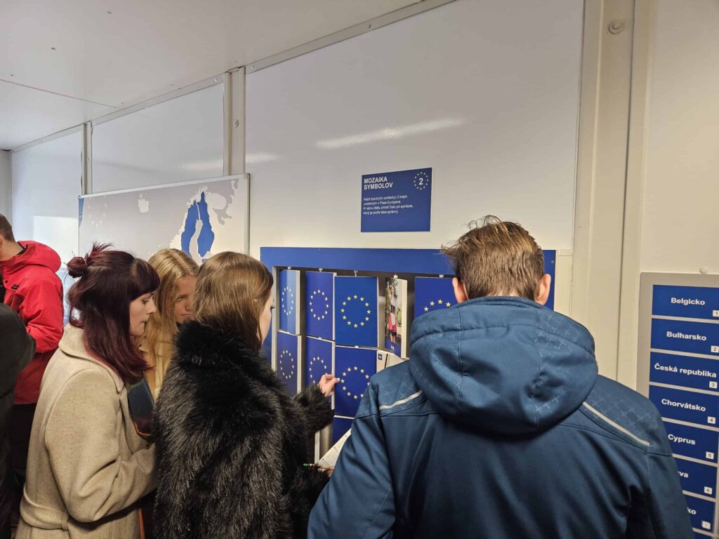 Niekoľko ľudí stojí pred nástenným displejom s malými modrými priehradkami s vlajkami EÚ a názvami krajín, komunikuje s displejom a pozerá sa do otvorených priehradiek.